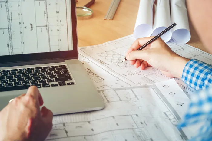 Händer som ritar på utskrivna CAD-ritningar vid en laptop
